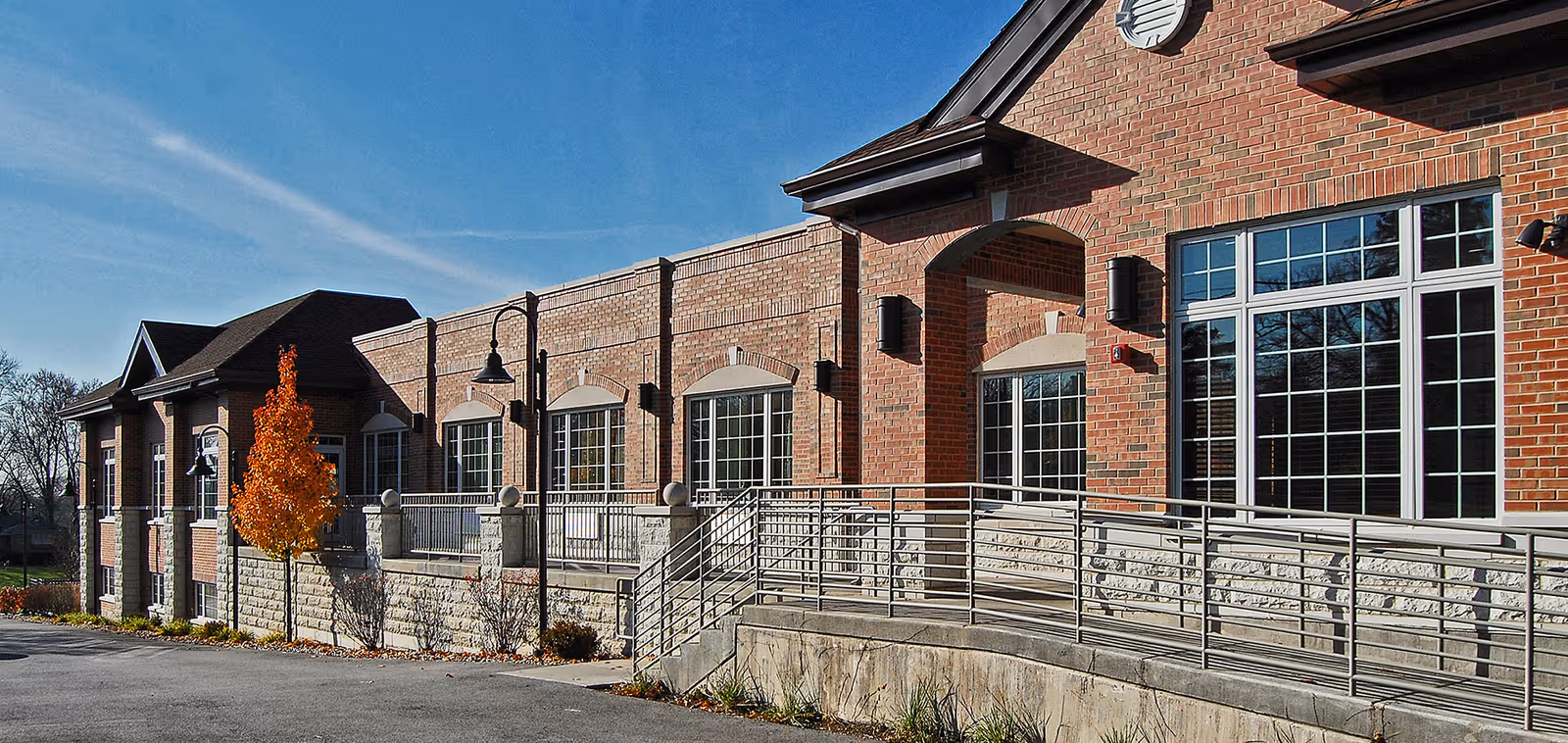 Exterior view of a brick building with large windows, a ramp with metal railings, and a small tree with orange leaves in front under a clear blue sky.