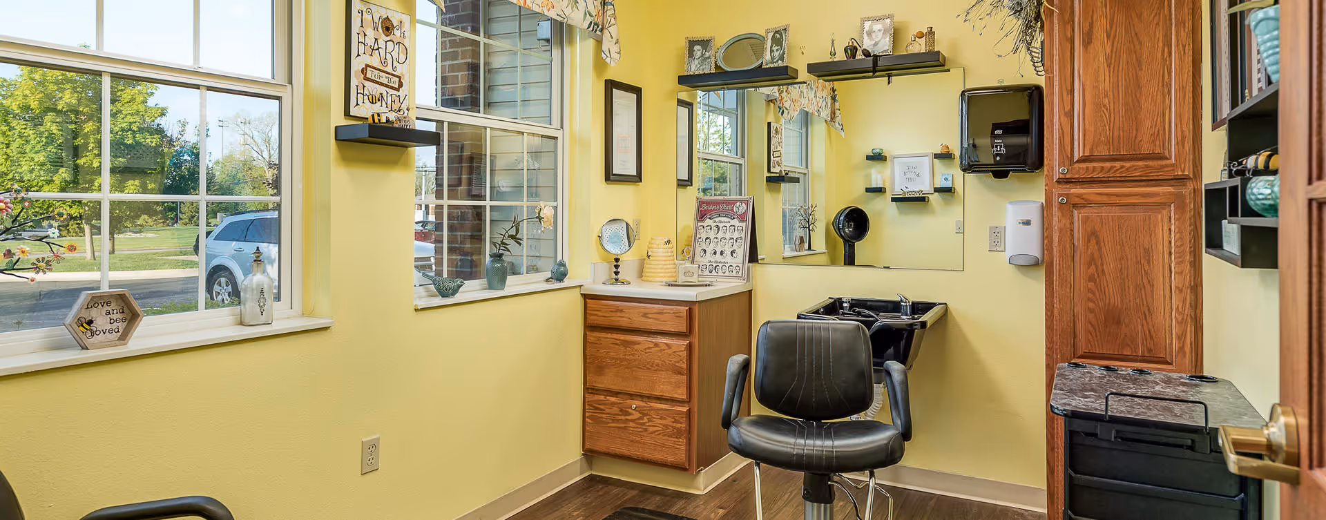 Interior view of a small salon or grooming room with yellow walls, a black salon chair, a sink for washing hair, a large mirror, wooden cabinets, and windows showing a green outdoor area with a parked car.
