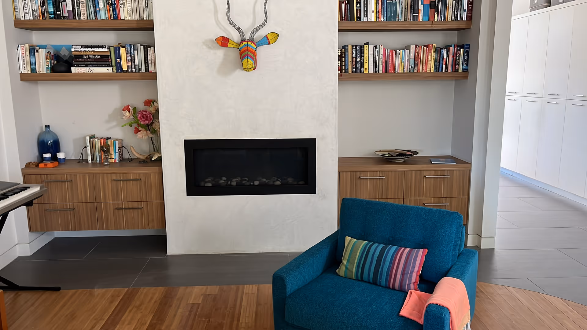 A cozy living room with a modern fireplace centered on a white textured wall. Above the fireplace is a colorful decorative antelope head. On either side of the fireplace are wooden shelves filled with books and cabinets below. A blue armchair with a striped cushion and a coral throw blanket is positioned in front of the fireplace. To the left, a keyboard is partially visible. The floor is a mix of wood and gray tiles, and a hallway with white cabinets is visible in the background.