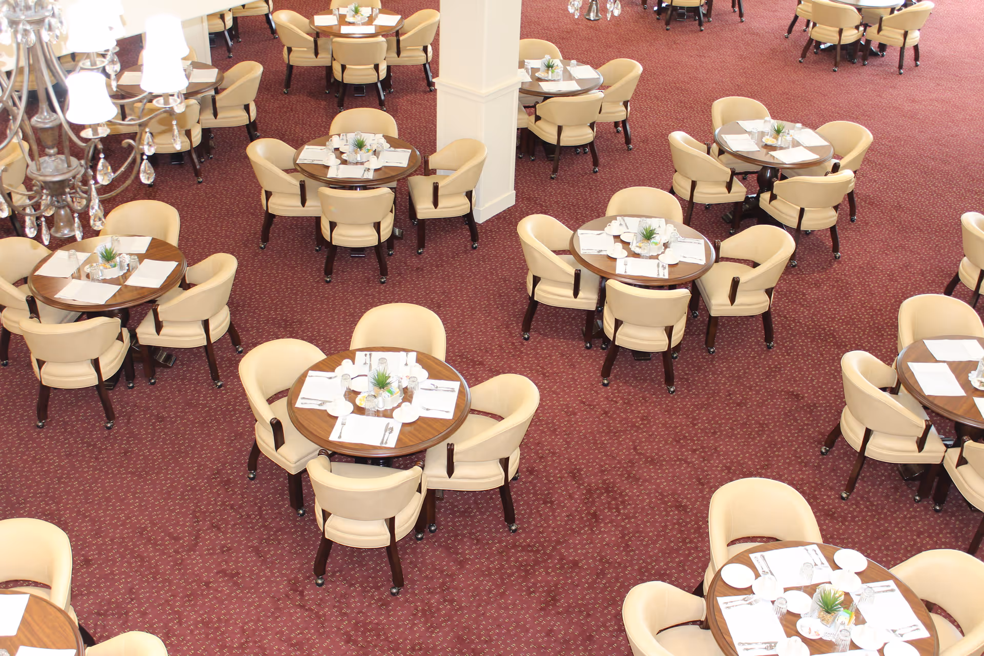 Overhead view of a dining room with multiple round tables, each surrounded by four beige upholstered chairs. The tables are set with placemats, napkins, utensils, and small decorative plants. The floor is covered with a red patterned carpet, and a chandelier is partially visible on the left side.