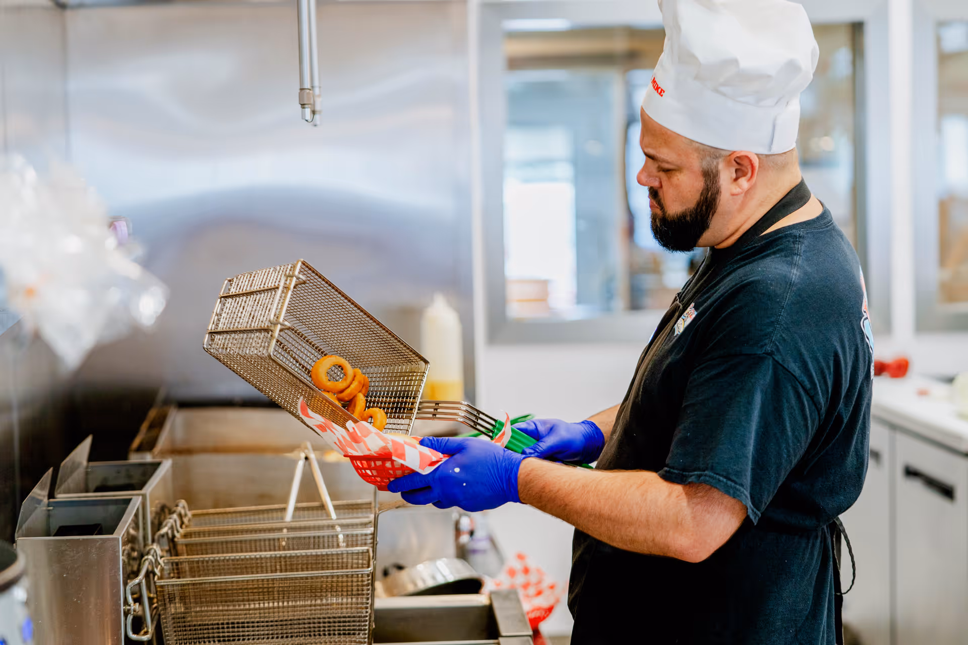 A cook wearing a white hat and blue gloves removes fried onion rings from a deep fryer into a paper basket in a commercial kitchen.