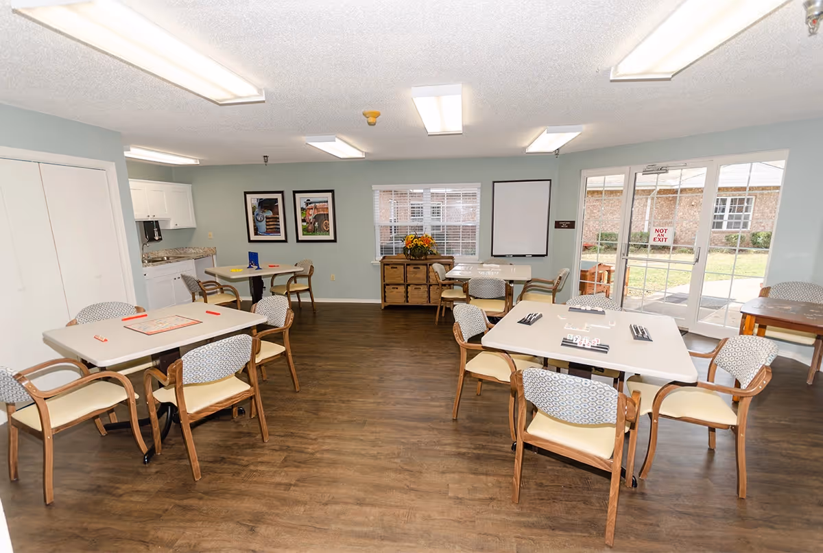 Bright communal activity/dining room with multiple tables and chairs, a small kitchenette, and glass doors leading to an outdoor courtyard.