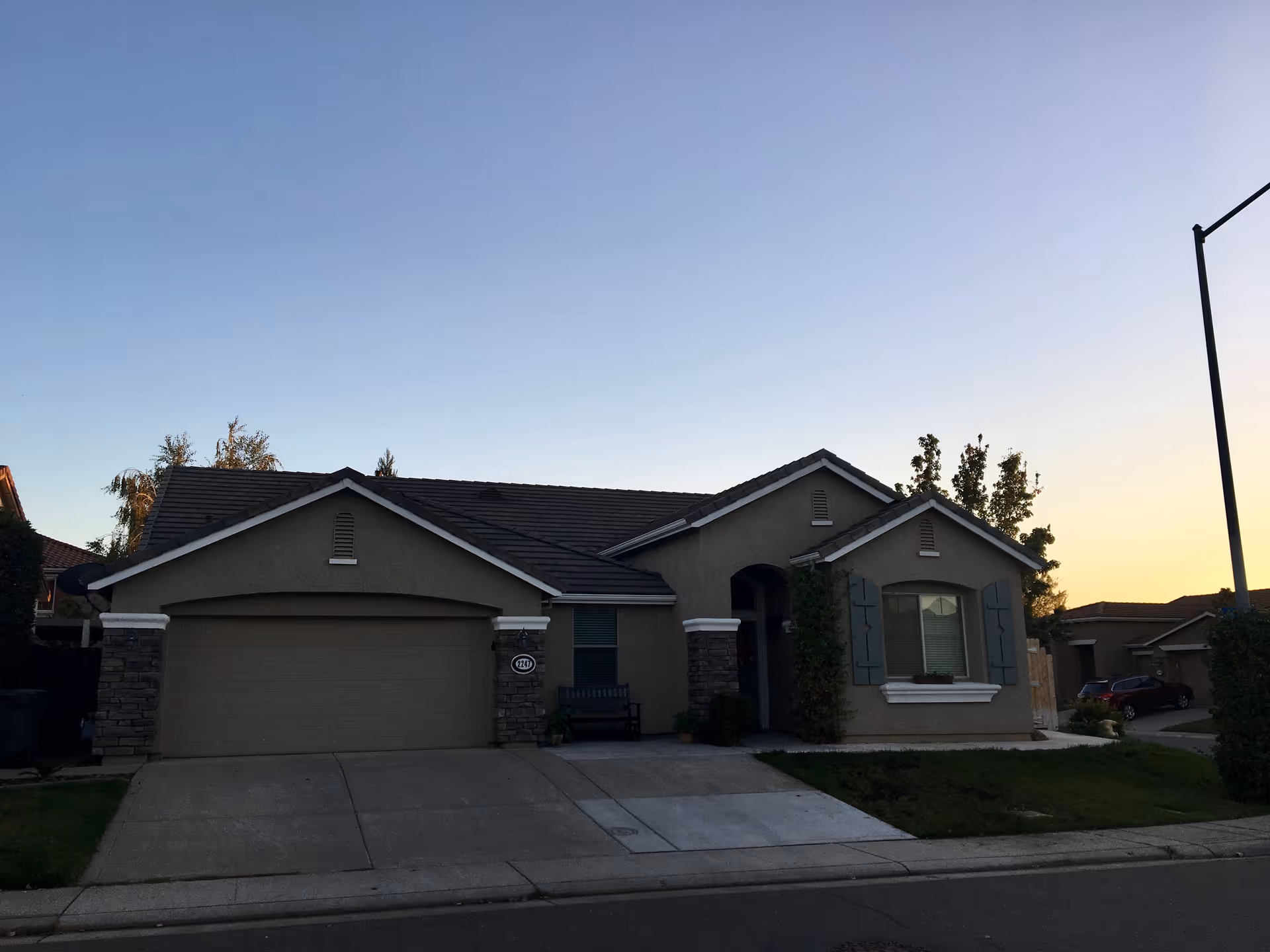 Exterior view of a single-story residential building with a two-car garage, a small front lawn, and a driveway at dusk. The sky is clear with a gradient from blue to light yellow near the horizon.