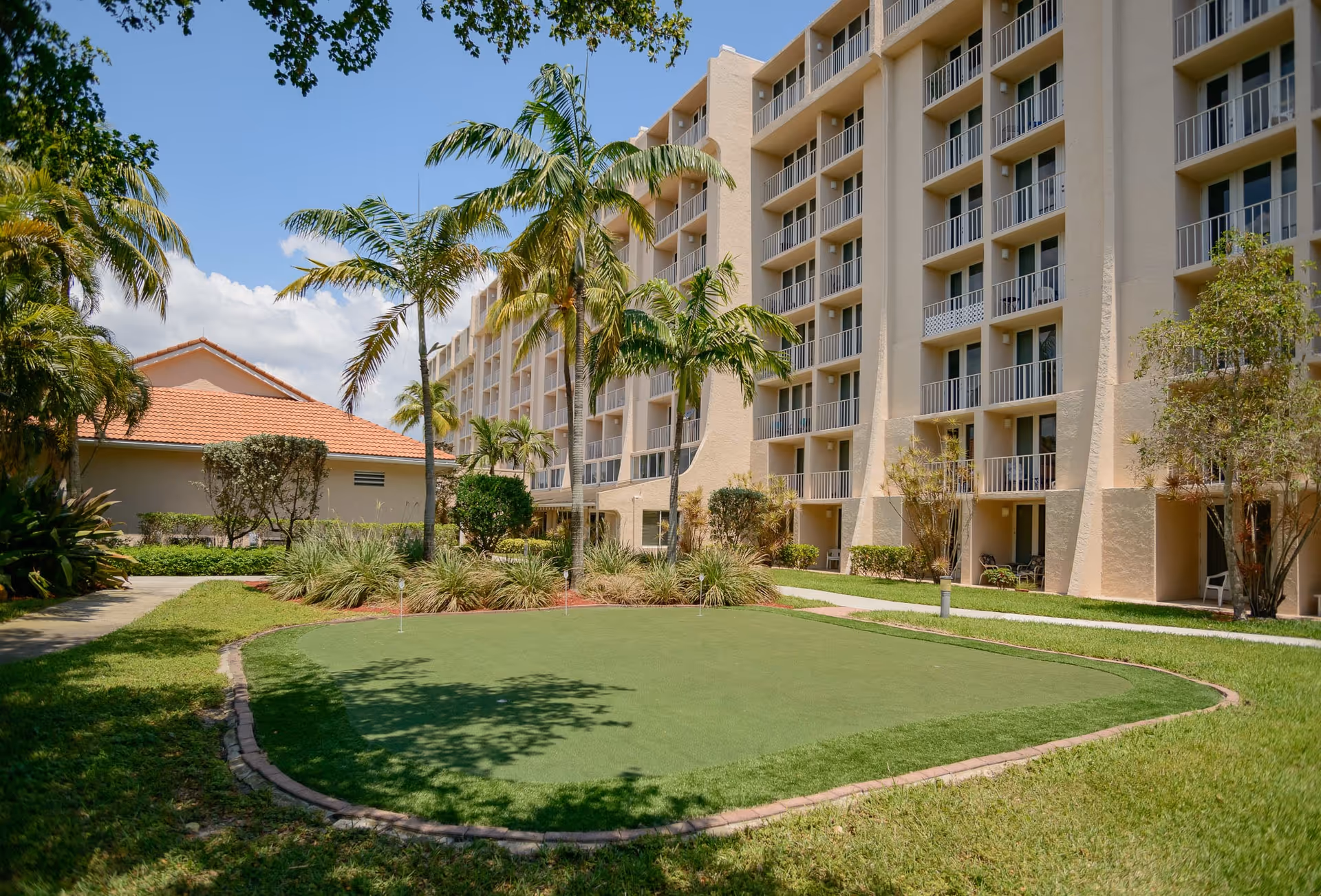 Outdoor view of a senior living facility with a putting green surrounded by grass, palm trees, and shrubbery. The building has multiple floors with balconies and beige exterior walls under a clear blue sky.