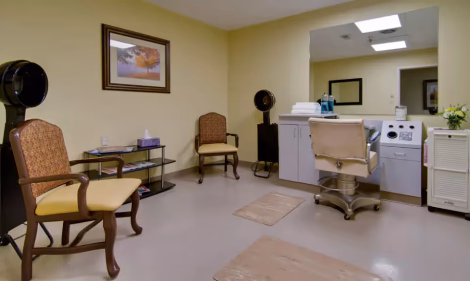 A small salon room with two vintage hair dryer chairs, two wooden chairs with yellow cushions, a small glass shelf with magazines and tissues, a large mirror above a counter with towels and hair care products, and beige walls with a framed picture of a tree.