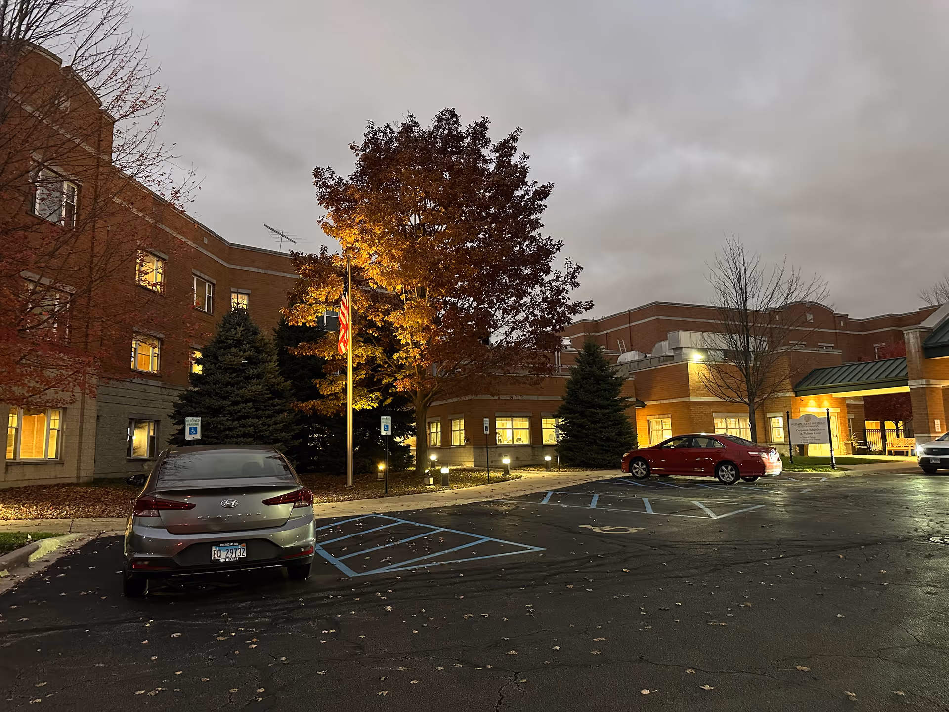 Exterior view of St. Joseph Village of Chicago at dusk, showing a brick building with lit windows, a parking lot with several cars, a flagpole with an American flag, and trees with autumn leaves.