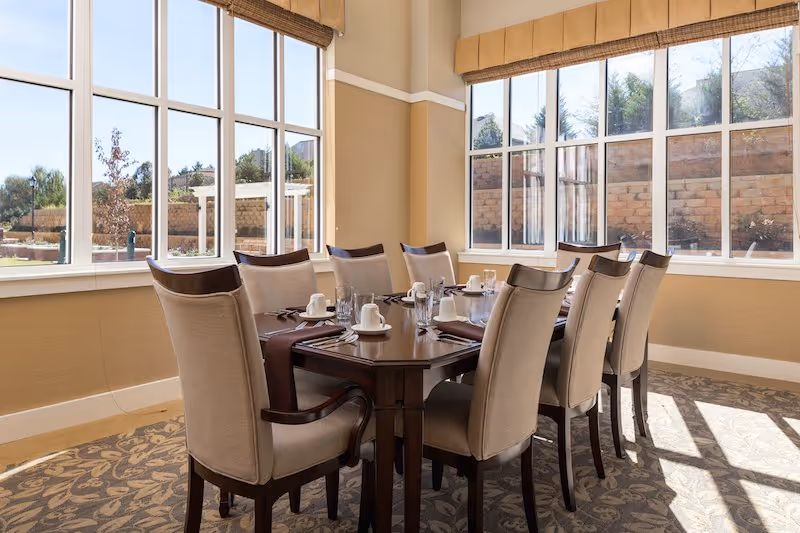 A bright dining room with a large rectangular wooden table set for eight people. The table is arranged with white cups, saucers, glasses, and silverware on brown placemats. The room has large windows on two sides allowing natural light to fill the space, with beige walls and patterned carpet flooring.