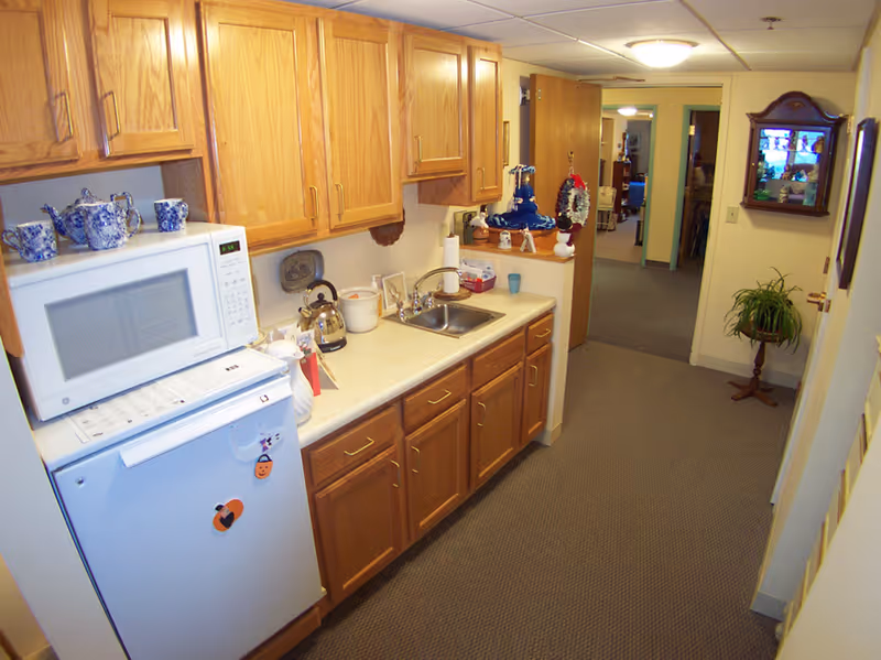 Small kitchenette with oak cabinets, a microwave over a mini-fridge, sink and countertop items along a carpeted hallway.
