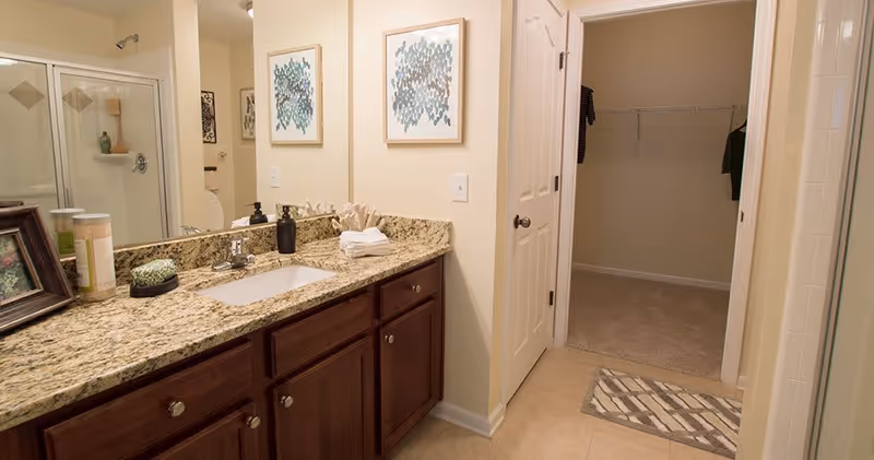 Bathroom with a granite countertop vanity featuring a sink, soap dispenser, and decorative items. There is a large mirror above the vanity, a shower with glass doors on the left, and an open doorway leading to a walk-in closet with hanging clothes. The walls are light-colored and the floor is tiled.