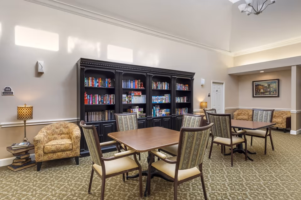 A cozy common area with two wooden tables surrounded by cushioned chairs, a large black bookshelf filled with books and board games against the wall, two patterned armchairs, two table lamps with patterned shades, and a framed painting on the far wall.