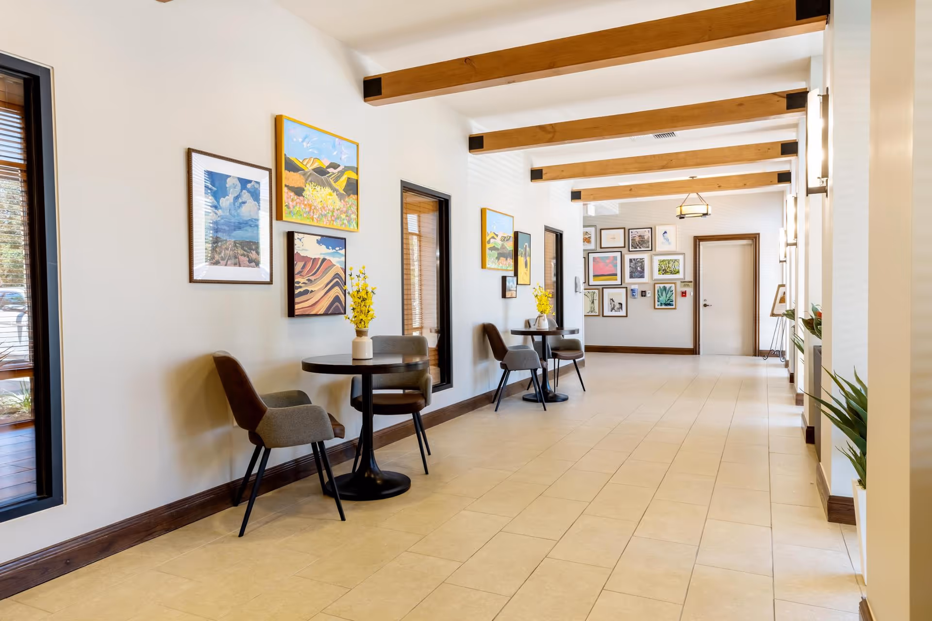 Bright interior hallway with small round tables and chairs, framed artwork on the walls, and exposed wooden ceiling beams.