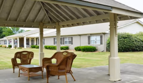 Outdoor covered seating area with wicker chairs and a small table on a concrete patio, with a single-story building and green lawn in the background.