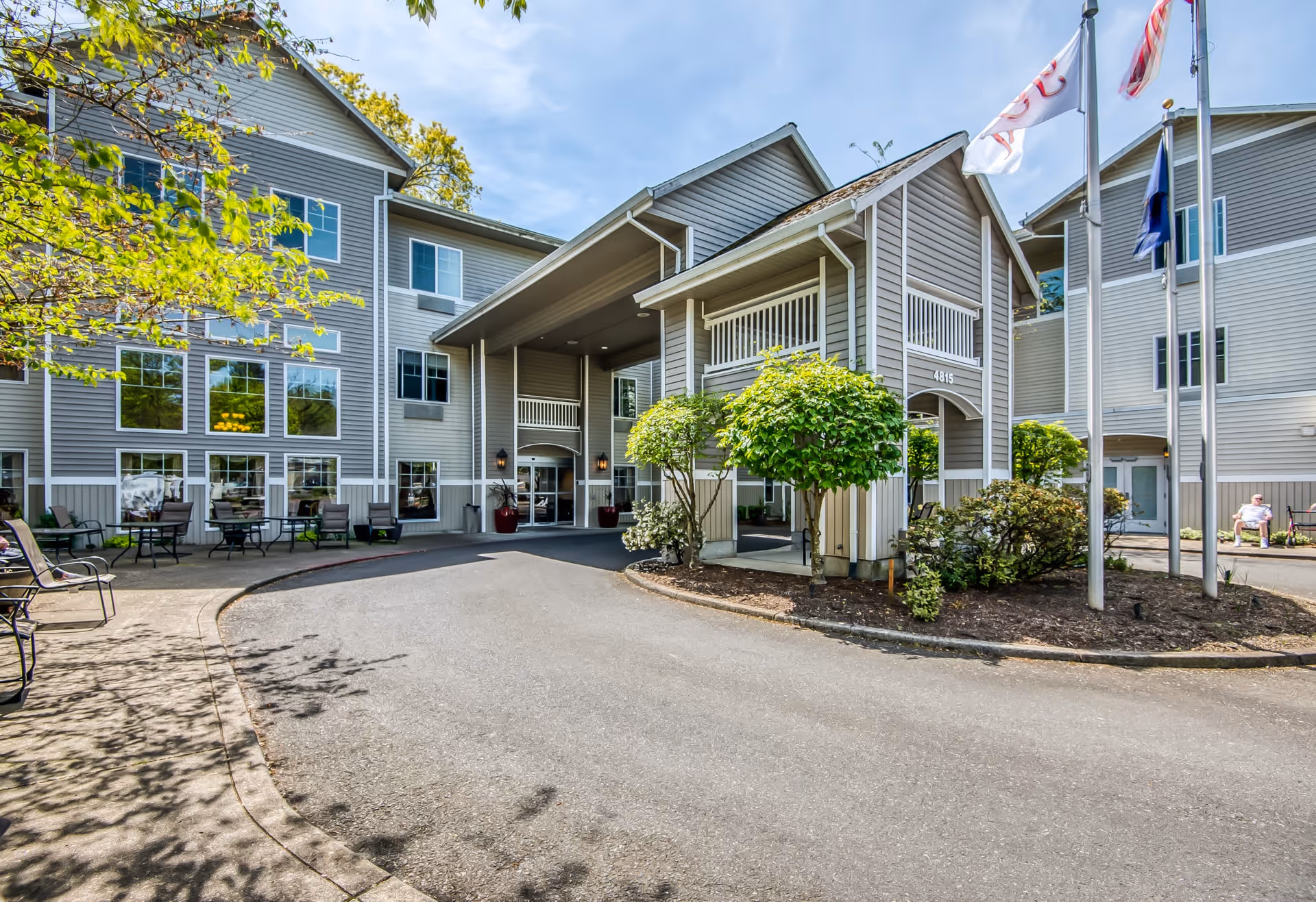 Exterior view of a multi-story assisted living facility with gray siding, large windows, and a covered entrance. There are small trees and shrubs planted near the entrance, flagpoles with flags, and outdoor seating areas with tables and chairs on the left side. A person is sitting on a bench near the right side of the building.