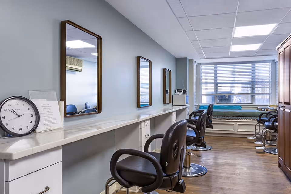 Interior view of a salon area with multiple black salon chairs in front of a long white countertop with mirrors mounted on the light blue wall. A clock and some papers are on the countertop. There is a window with blinds at the far end and a cushioned bench beneath it. The floor is wooden and the ceiling has white tiles.