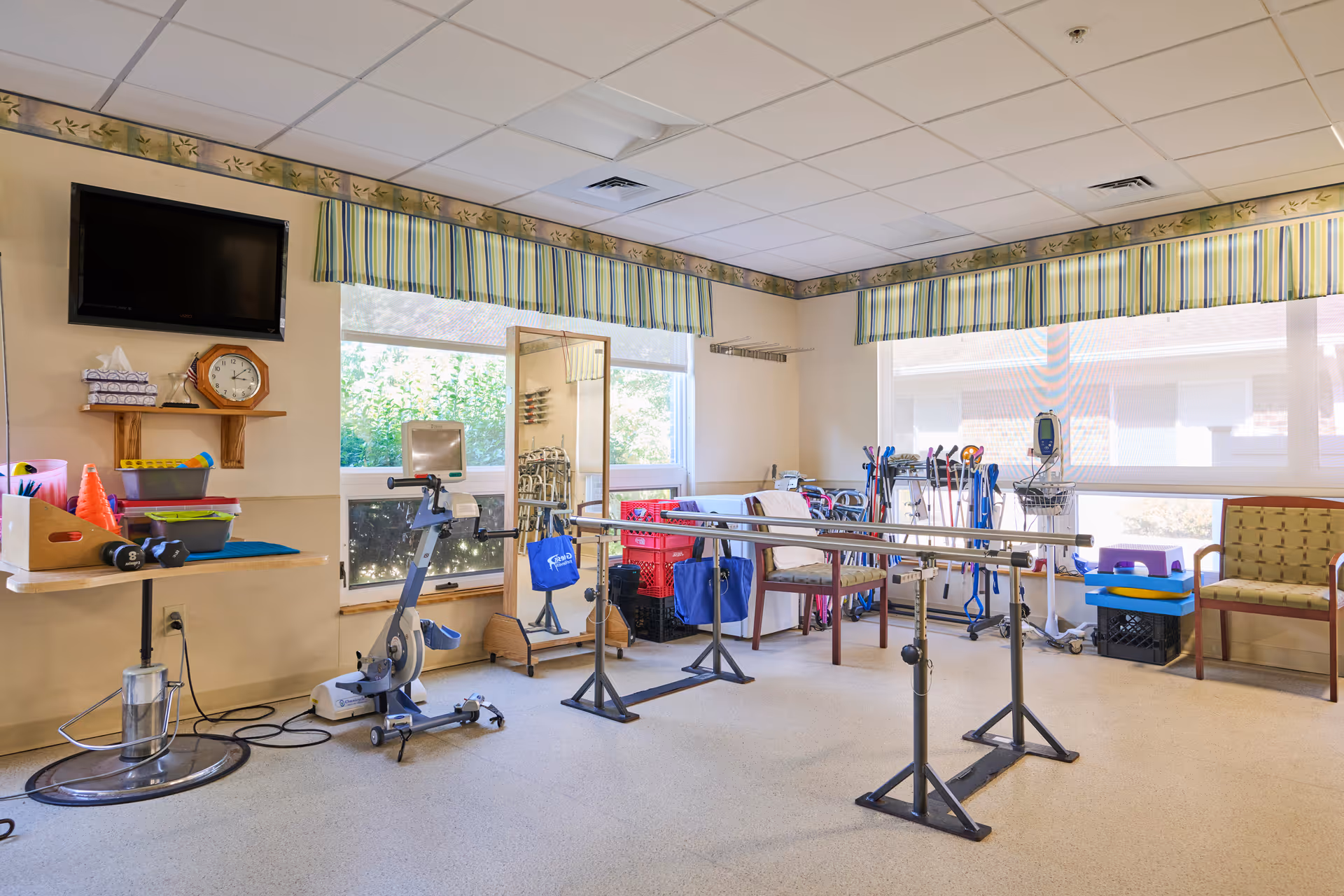 A rehabilitation therapy room with parallel bars for walking exercises, a stationary exercise bike, a full-length mirror, various mobility aids like walkers and canes, chairs, and a TV mounted on the wall. The room has large windows with striped valances allowing natural light to enter.
