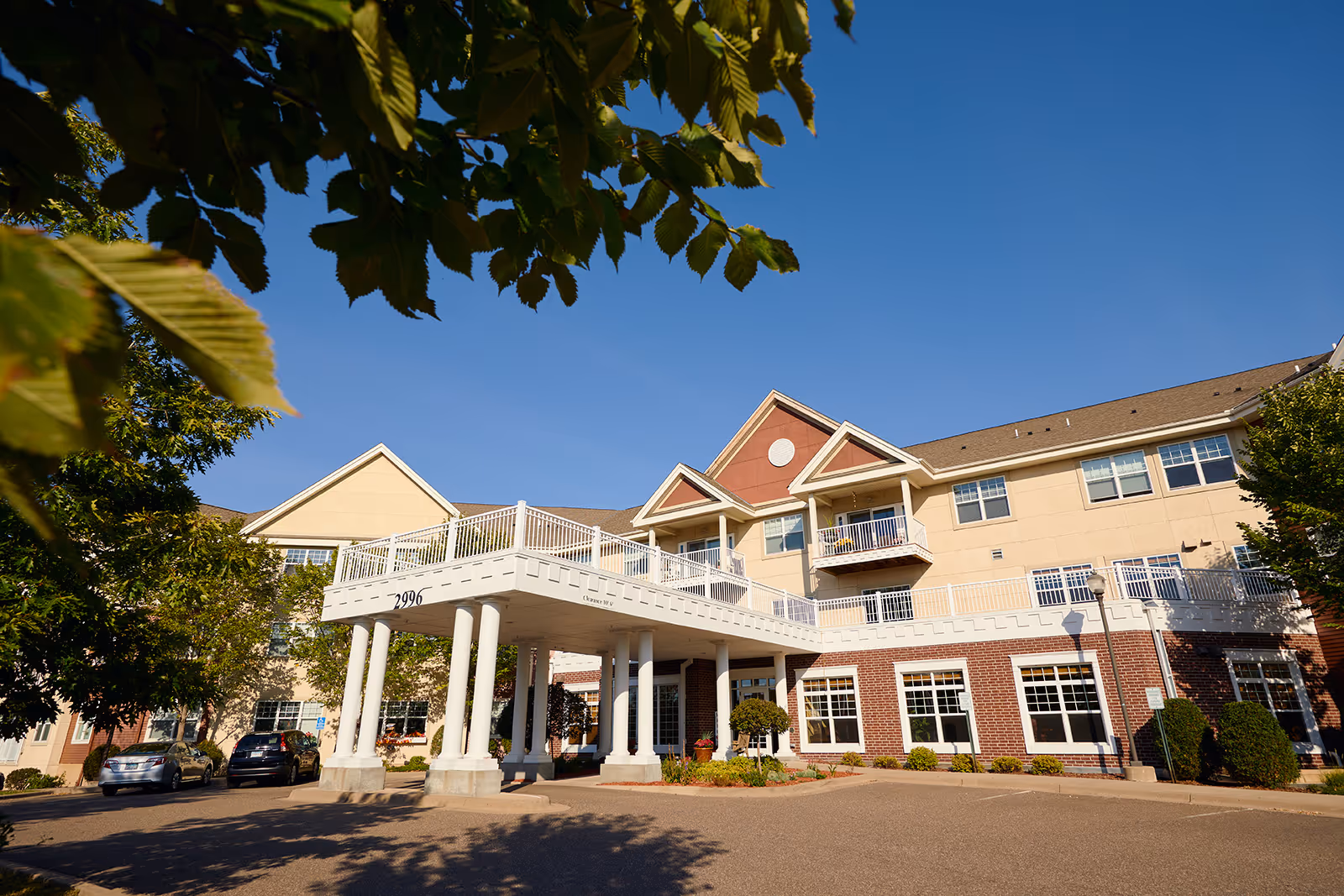 Front entrance of a three-story assisted living building with a columned porte-cochère, balconies, and landscaped trees under a clear blue sky.
