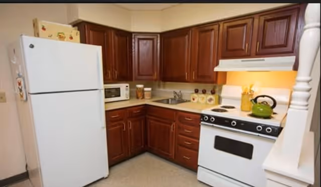 A compact kitchen with dark wooden cabinets, a white refrigerator, a microwave on the countertop, a sink, and a white stove with a green kettle on top. The kitchen has a warm, cozy feel with under-cabinet lighting illuminating the stove area.