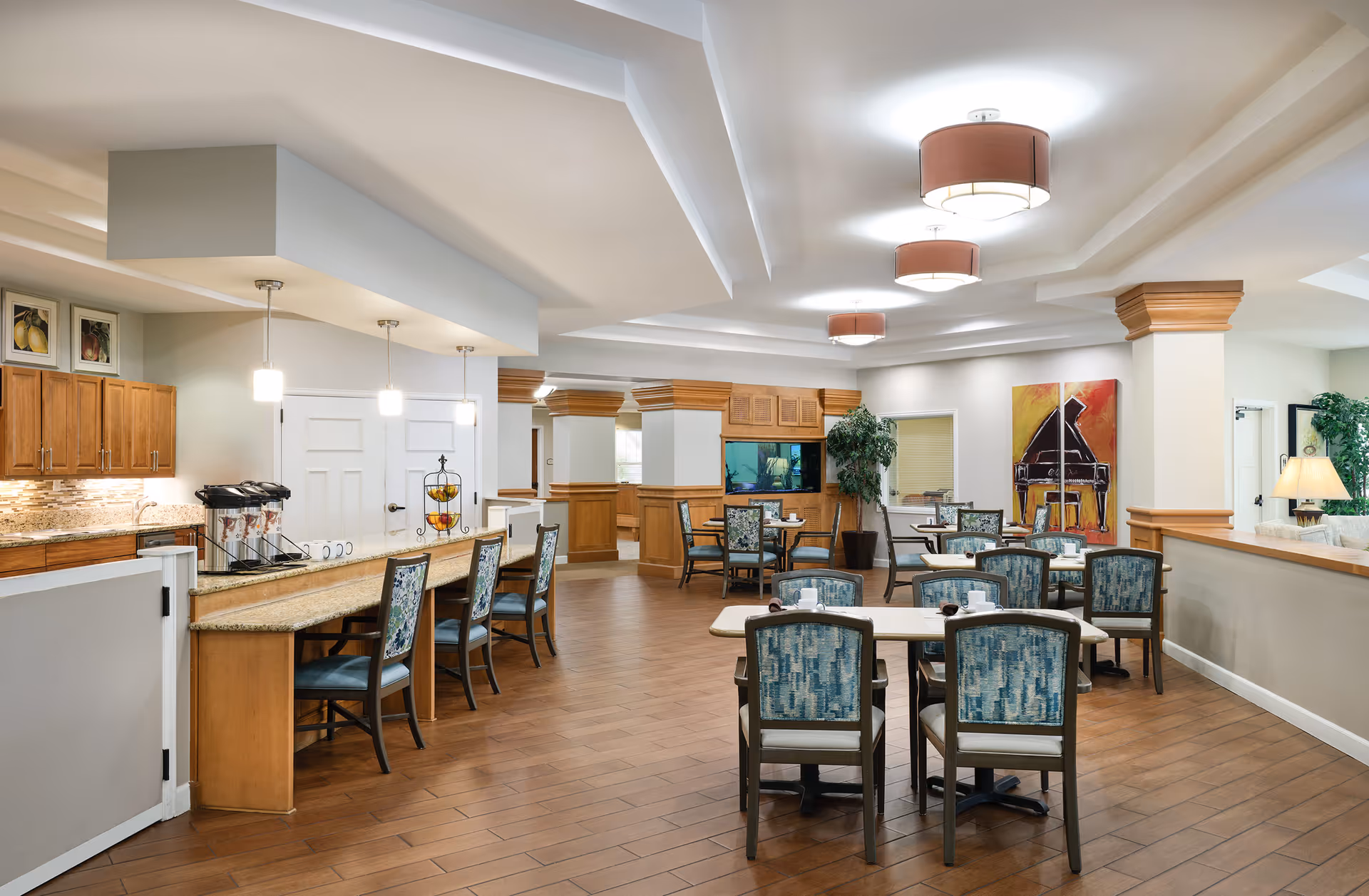 A spacious dining area in a senior living facility with multiple tables and chairs arranged neatly. The room features wooden cabinetry, a countertop with bar stools, pendant lights, and ceiling fixtures. There is a large colorful painting of a piano on the wall, a fish tank built into the cabinetry, and some potted plants adding greenery to the space.