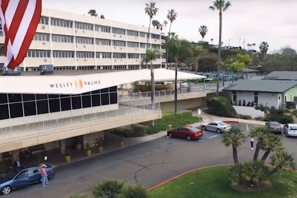 Exterior view of Wesley Palms Retirement Community building with multiple floors, palm trees, parked cars, and a few people near the entrance.