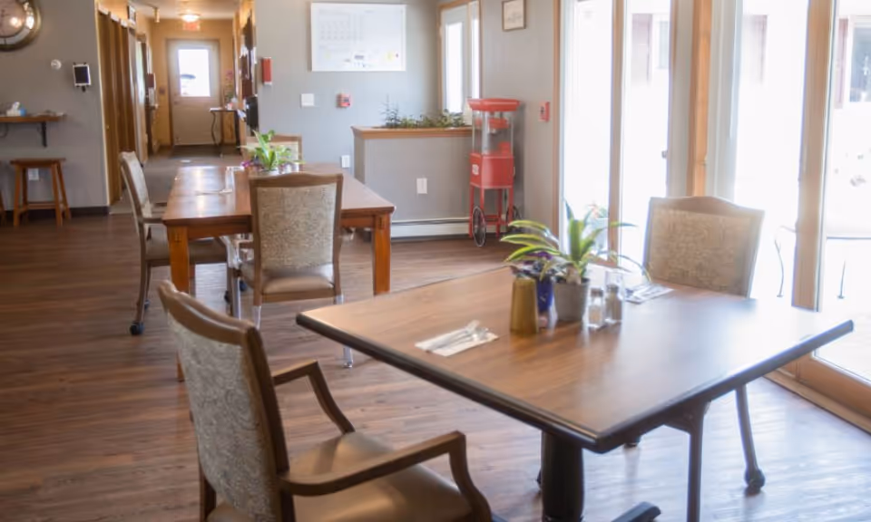 A bright dining area with wooden floors and tables set with plants, salt and pepper shakers, and utensils. There are upholstered chairs around the tables, large windows letting in natural light, and a red popcorn machine in the background near a wall with a calendar.