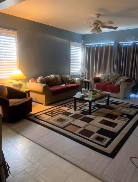 Living room with two sofas and an armchair around a coffee table on a geometric rug, lamps, and closed curtains.