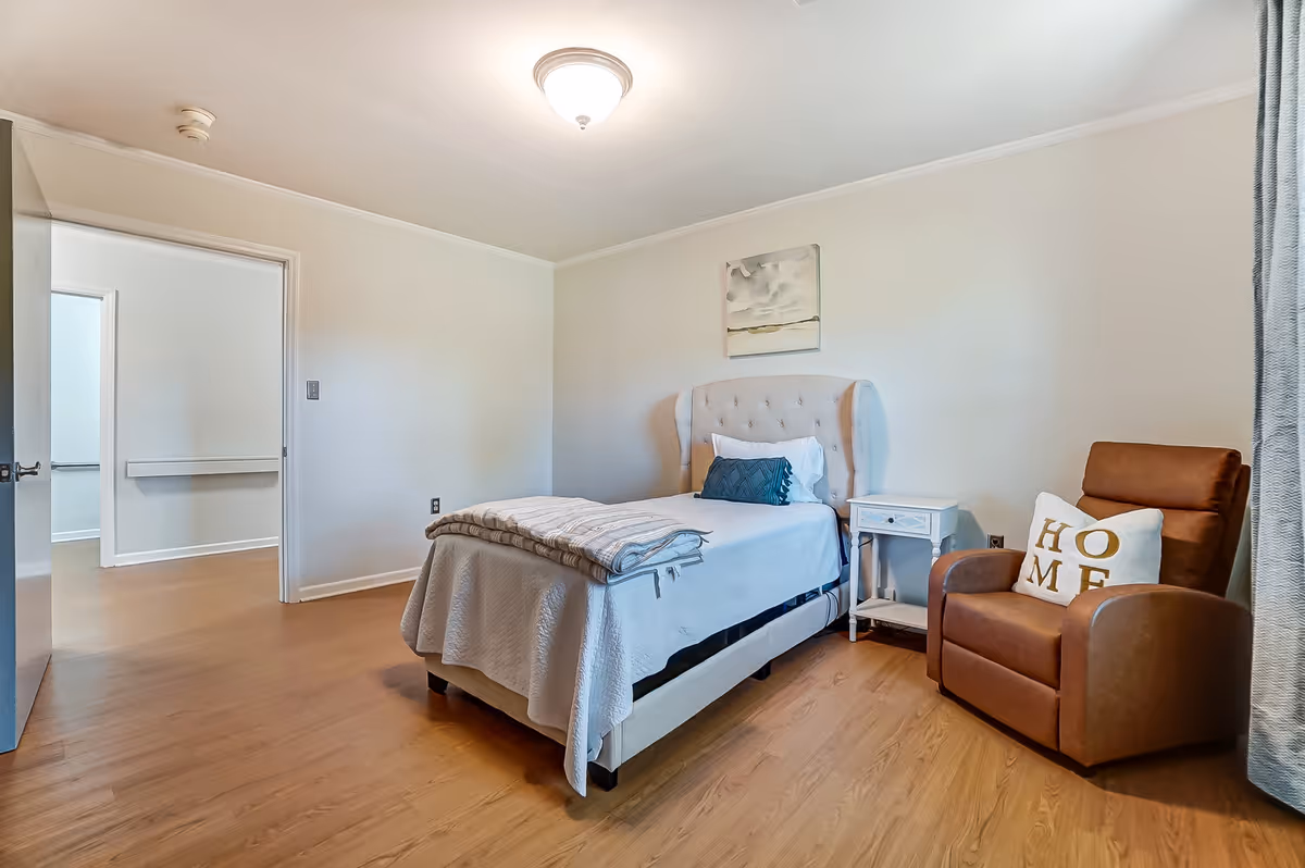 A simple bedroom with a single bed covered in a white quilt and a folded blanket at the foot. There is a white nightstand next to the bed and a brown recliner chair with a white pillow that has the word 'HOME' on it. The room has light-colored walls, wooden flooring, a ceiling light fixture, and a painting above the bed. A doorway leads to another room with similar flooring and walls.