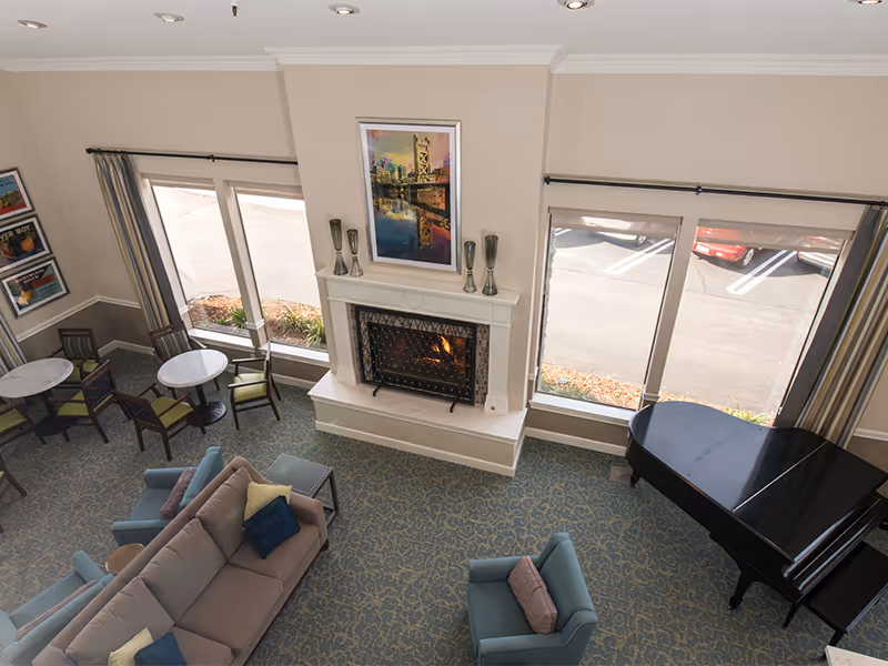 A bright communal lounge with a fireplace, sofas and chairs, round tables, and a grand piano seen from above.