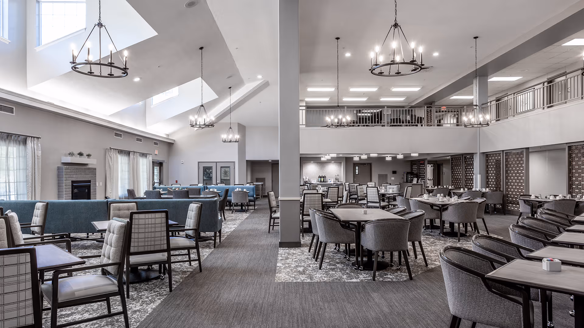 Spacious dining area with multiple tables and chairs arranged neatly. The room features high ceilings with large windows allowing natural light to fill the space. There are modern chandeliers hanging from the ceiling and a fireplace on the left side. The area has a mix of cushioned armchairs and dining chairs, with a carpeted floor and a second-floor balcony overlooking the room.