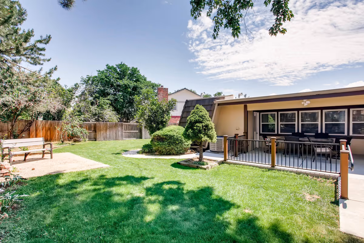 Sunny grassy courtyard with a patio seating area, bench, and surrounding trees beside a single-story building.