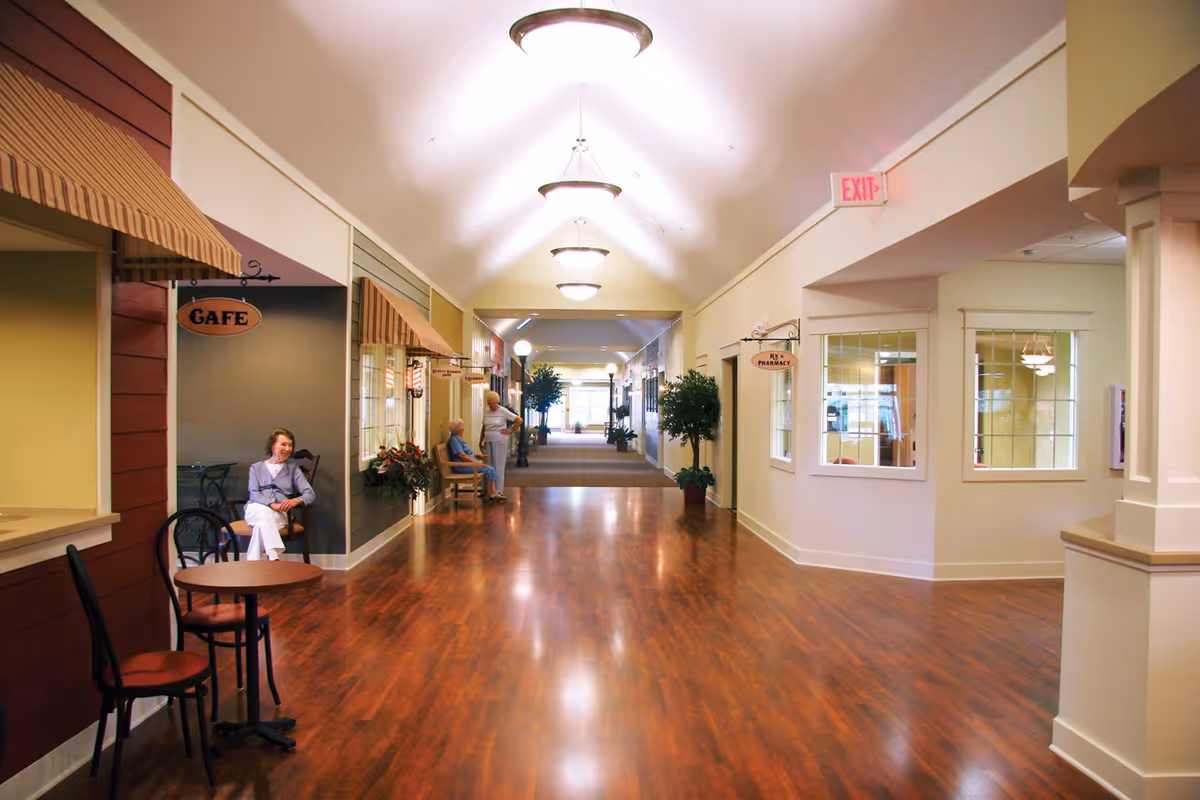 A wide, well-lit hallway in a senior living facility with wooden flooring and ceiling lights. On the left side, there is a small cafe area with a table and chairs where a woman is sitting. Further down the hallway, two elderly people are seated on benches near potted plants. Signs for a cafe and a pharmacy are visible along the walls.