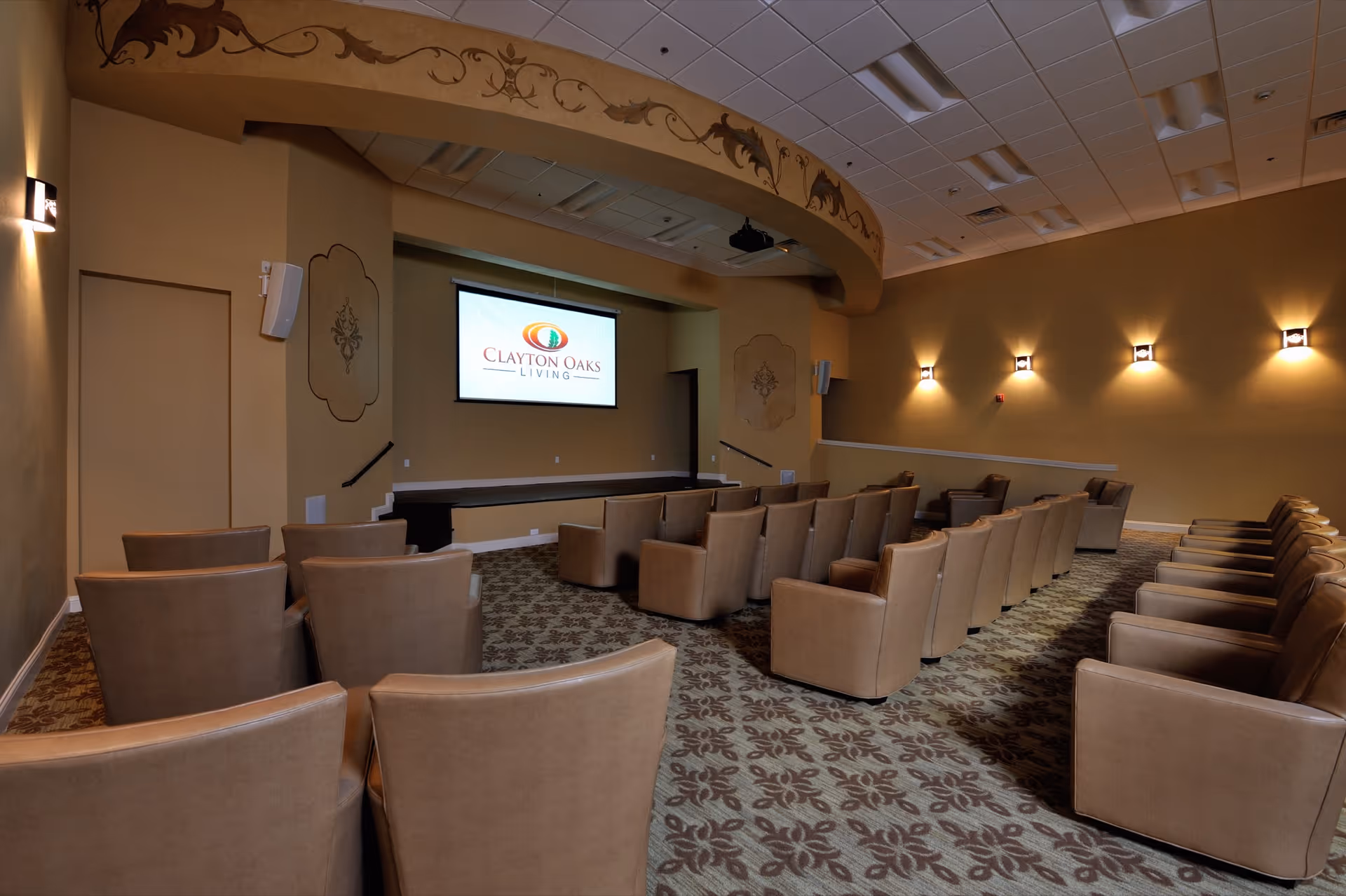 Small theater-style screening room with rows of tan upholstered chairs facing a projection screen displaying the Clayton Oaks Living logo.