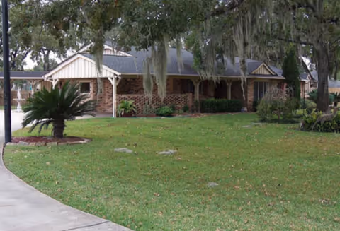 A single-story brick building with a covered porch surrounded by a well-maintained lawn and large trees draped with Spanish moss.