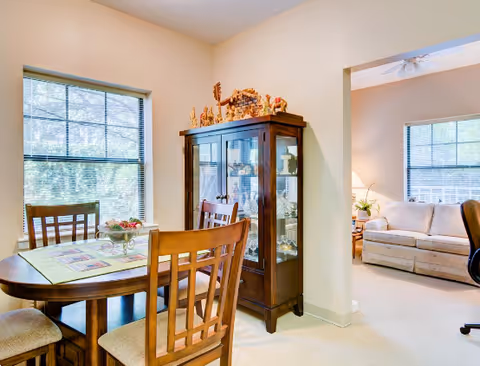 A bright dining area with a wooden table and four chairs, a glass-fronted cabinet displaying decorative items, and a window with blinds letting in natural light. Adjacent to the dining area is a living room with a beige sofa, a side table with a lamp, and a window with blinds.