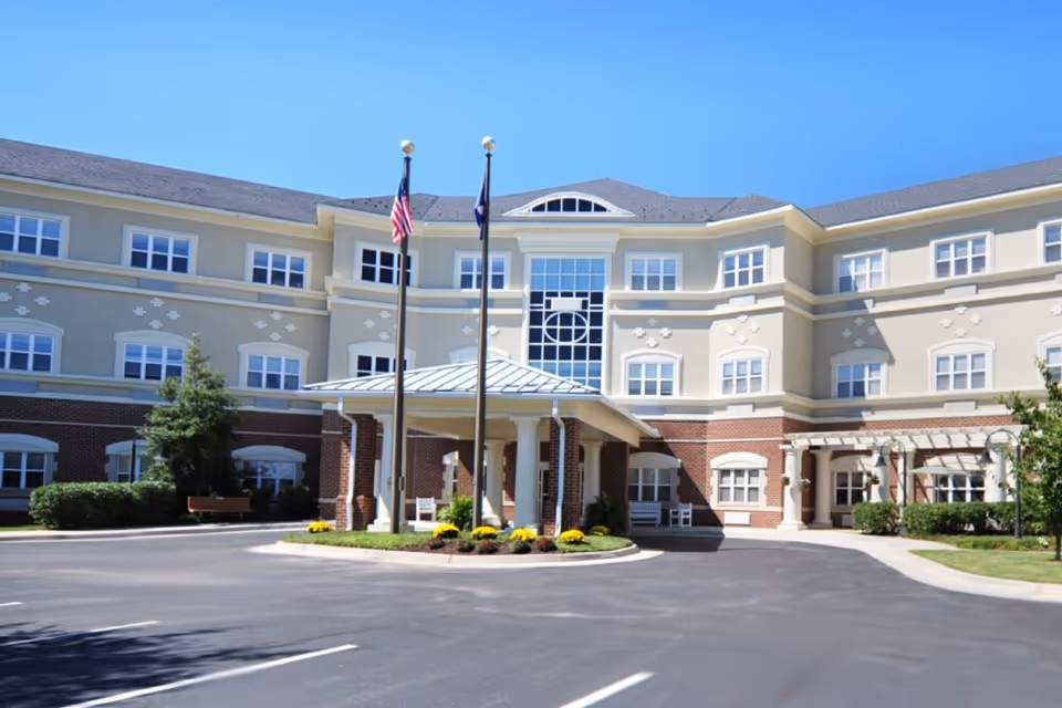 Exterior view of a three-story senior living facility building with beige and brick facade, multiple windows, a covered entrance with white columns, two flagpoles with flags, and a paved driveway in front under a clear blue sky.