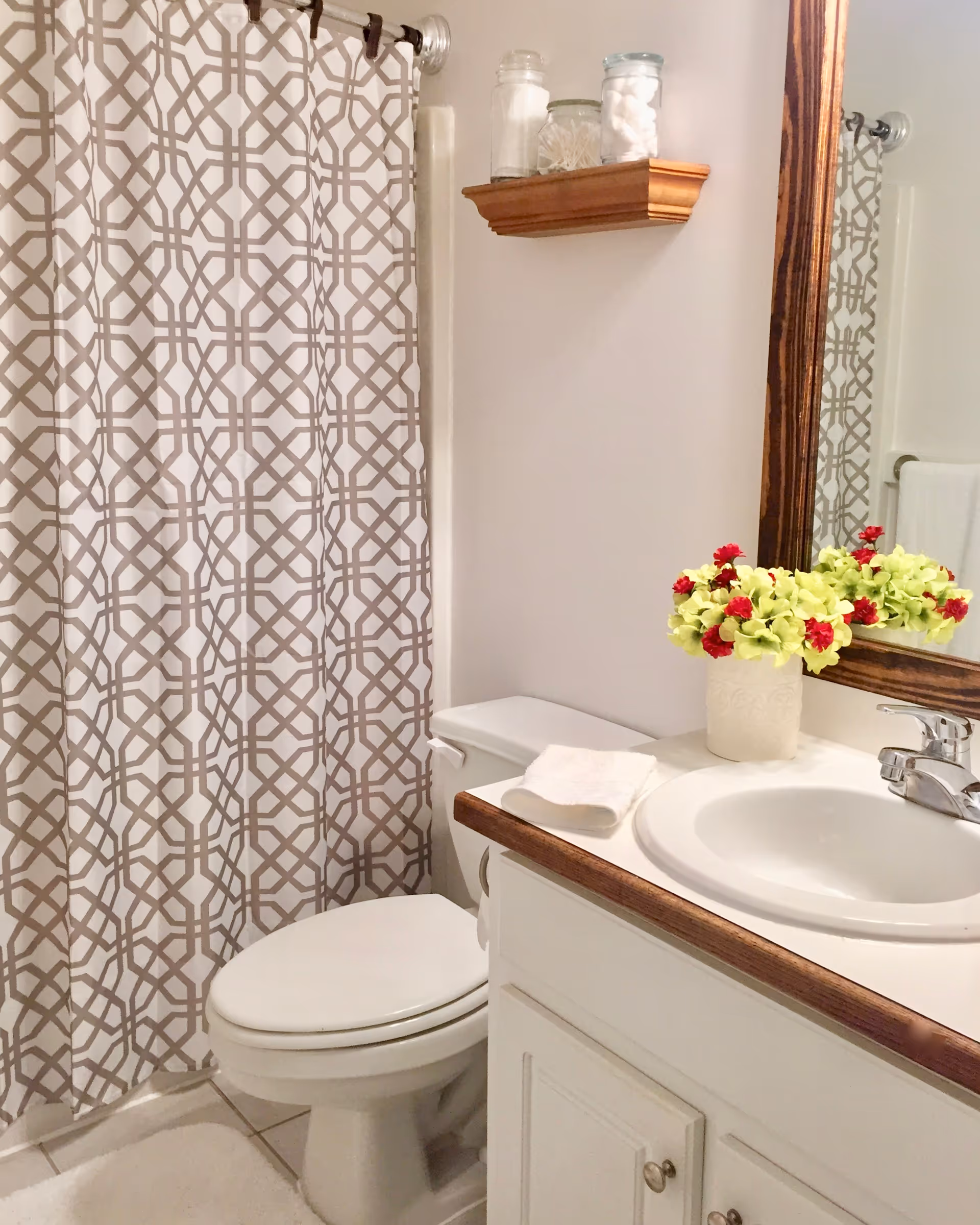 Small bathroom with a toilet, sink and vanity, patterned shower curtain, and a vase of flowers on the counter.