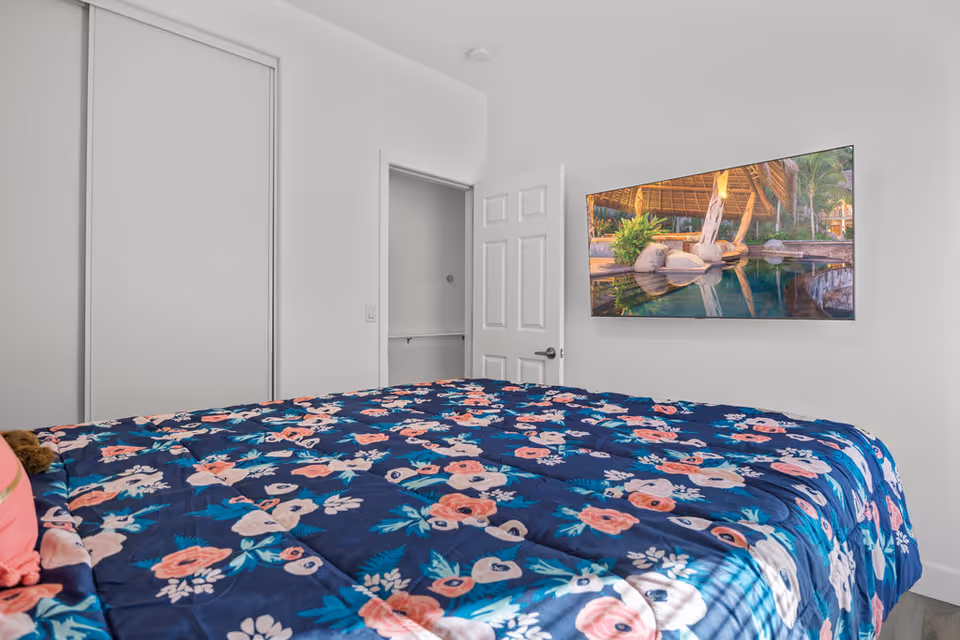 A bedroom with a bed covered in a blue floral comforter. The room has white walls, a closed closet door, an open door leading to another room, and a flat-screen TV mounted on the wall displaying an outdoor scene with water and greenery.