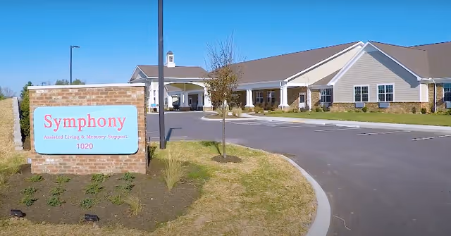 Exterior view of Symphony Assisted Living and Memory Support facility showing a single-story building with a covered entrance, a driveway, and a landscaped area with a sign that reads 'Symphony Assisted Living & Memory Support 1020'.