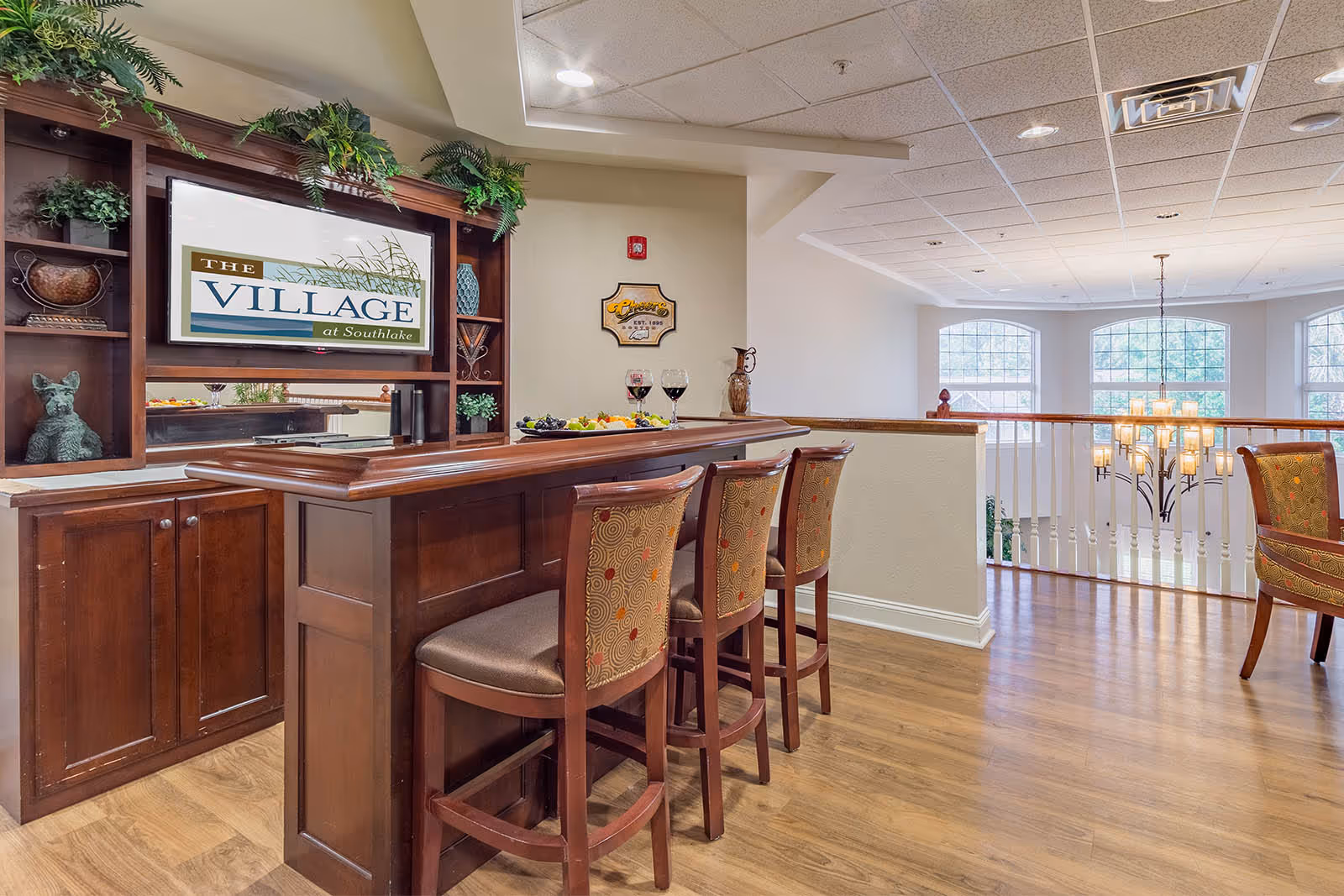 Interior common-area bar with a wooden counter and three upholstered bar stools overlooking a bright two-story atrium.