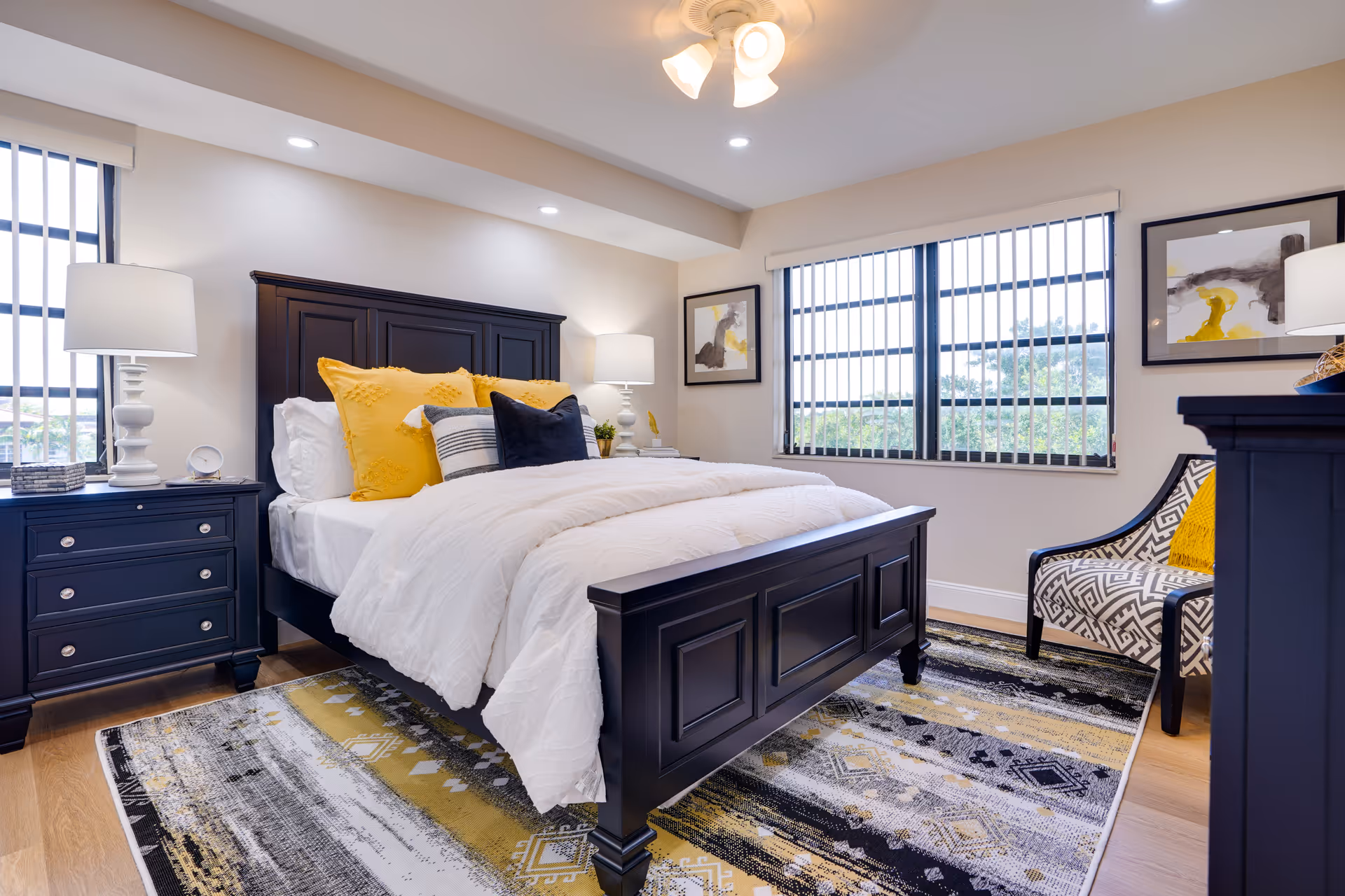 A bright and cozy bedroom featuring a dark wooden bed frame with white bedding and yellow and black accent pillows. The room has two windows with vertical blinds, two matching dark wooden nightstands with white lamps, a patterned armchair with a yellow pillow, framed abstract artwork on the walls, and a multicolored area rug with shades of yellow, black, and gray on a wooden floor.