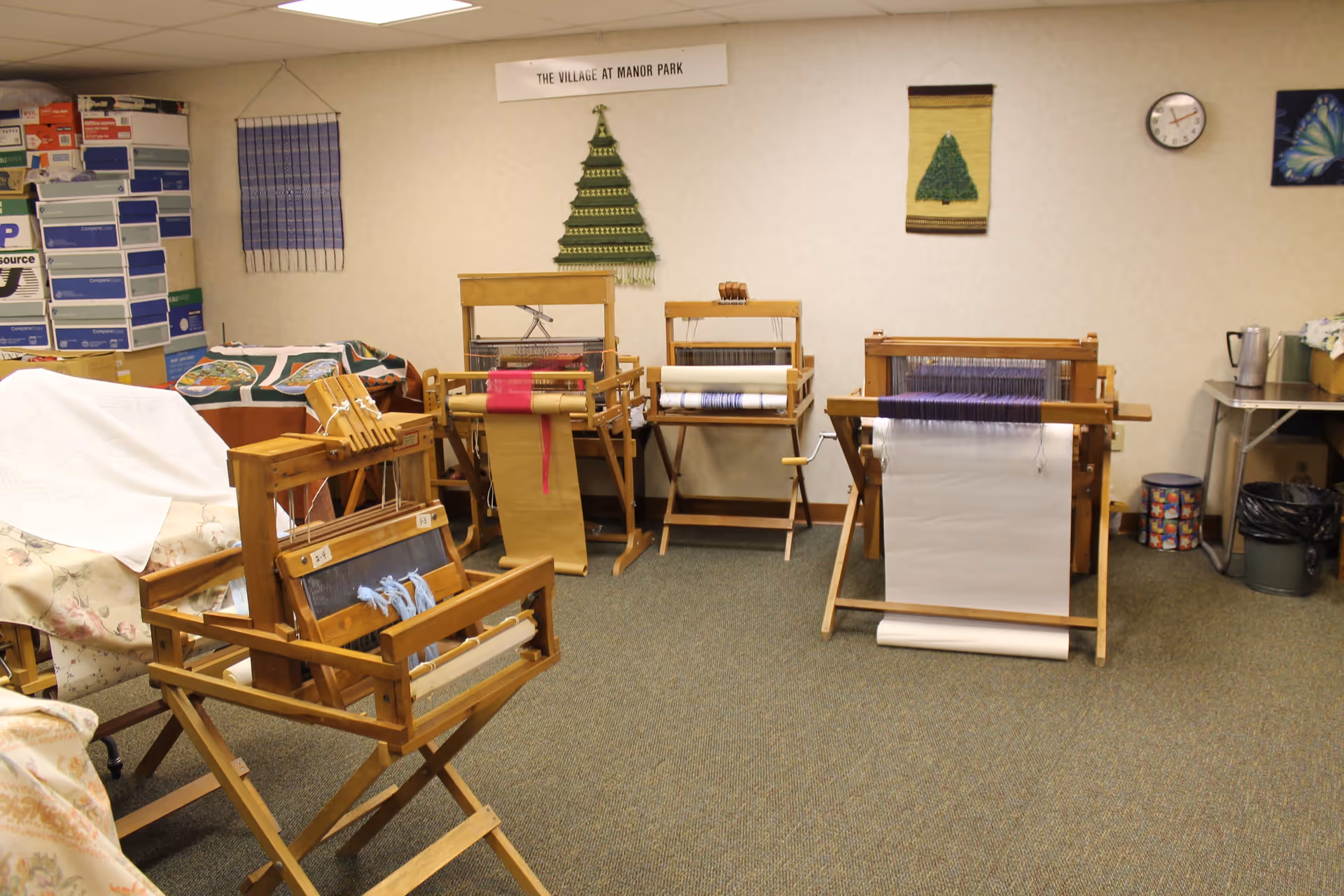 A room with several wooden weaving looms set up on a carpeted floor. The walls have decorative woven wall hangings, including two green tree designs and a butterfly picture. There are stacks of boxes and some covered furniture on the left side, and a clock on the wall on the right side. A sign on the wall reads 'THE VILLAGE AT MANOR PARK'.