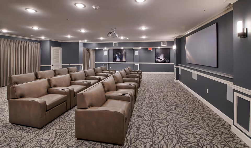 A modern theater room with multiple rows of brown leather recliner seats facing a large blank projection screen on a dark gray wall. The room has patterned carpet flooring, recessed ceiling lights, wall sconces, and framed artwork on the walls.