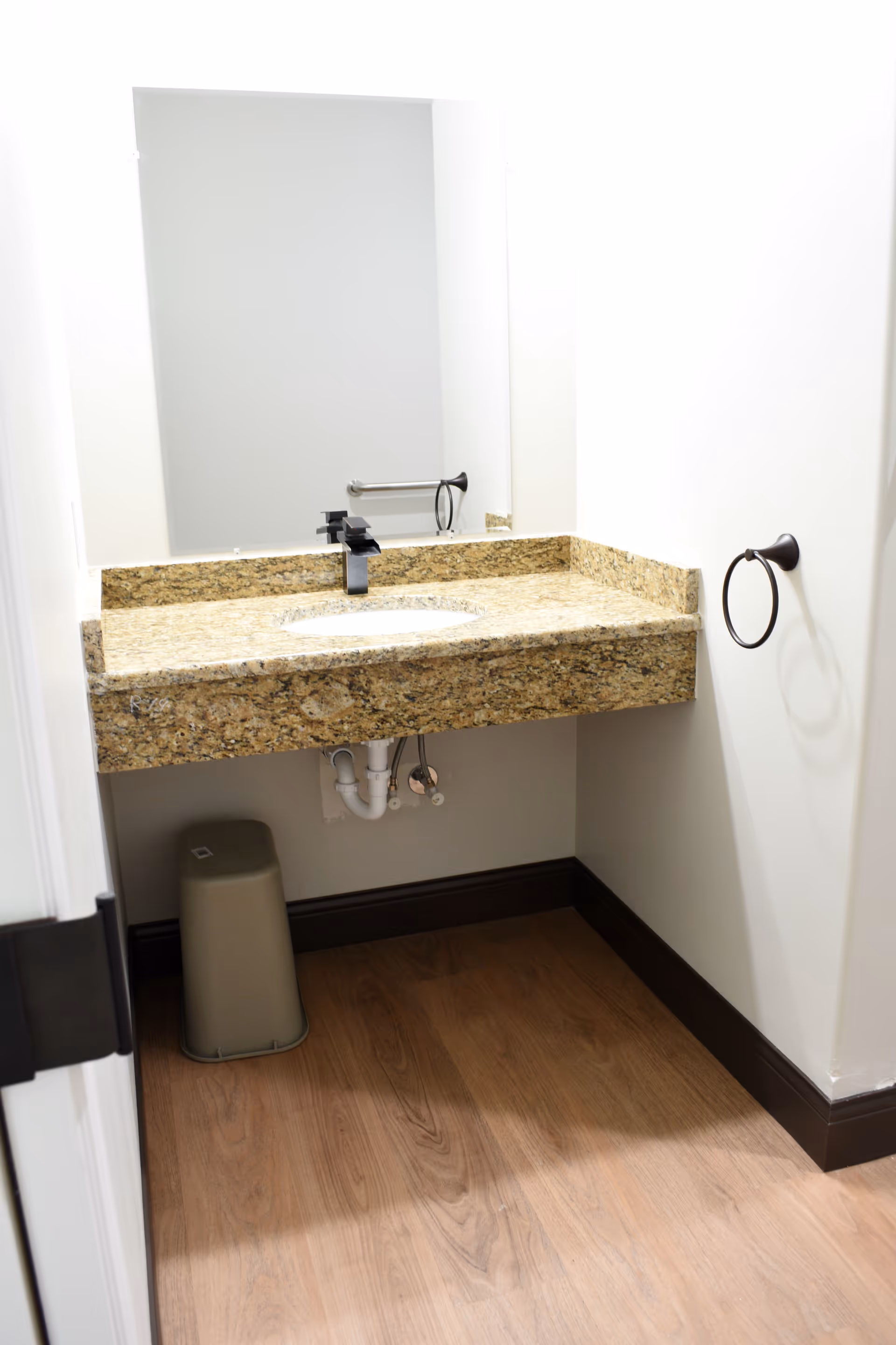 A bathroom sink area with a granite countertop, a black faucet, a large rectangular mirror above the sink, a towel ring mounted on the wall to the right, and a small trash bin placed under the sink. The floor is wooden and the walls are painted white.