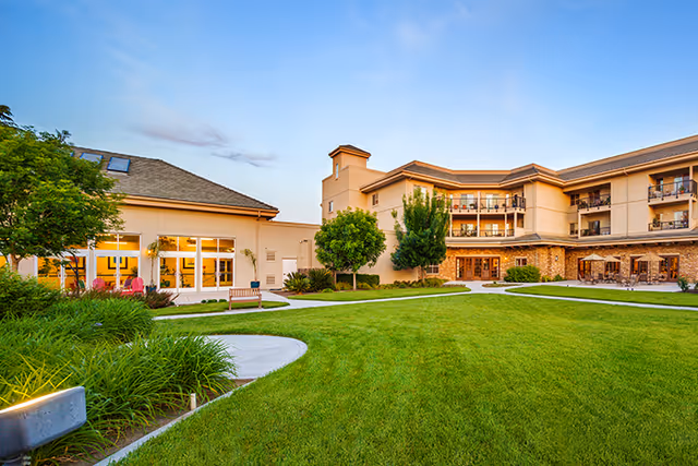 Exterior view of Covenant Living of Turlock facility showing a well-maintained green lawn, landscaped bushes, trees, and a multi-story building with balconies and large windows under a clear blue sky.