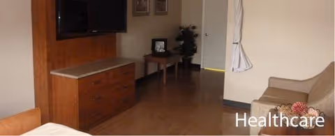 Interior view of a room with a wooden dresser topped with a flat-screen TV, a small table with a plant and picture frame, a beige armchair with a decorative pillow, and a door with a curtain hanging beside it.