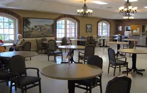 A spacious senior living dining hall with round tables, chairs, chandeliers, and arched windows.