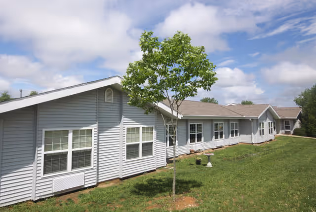 Exterior view of a single-story building with light gray siding and multiple white-framed windows under a partly cloudy sky. A small tree and a birdbath are visible on the grassy lawn in front of the building.