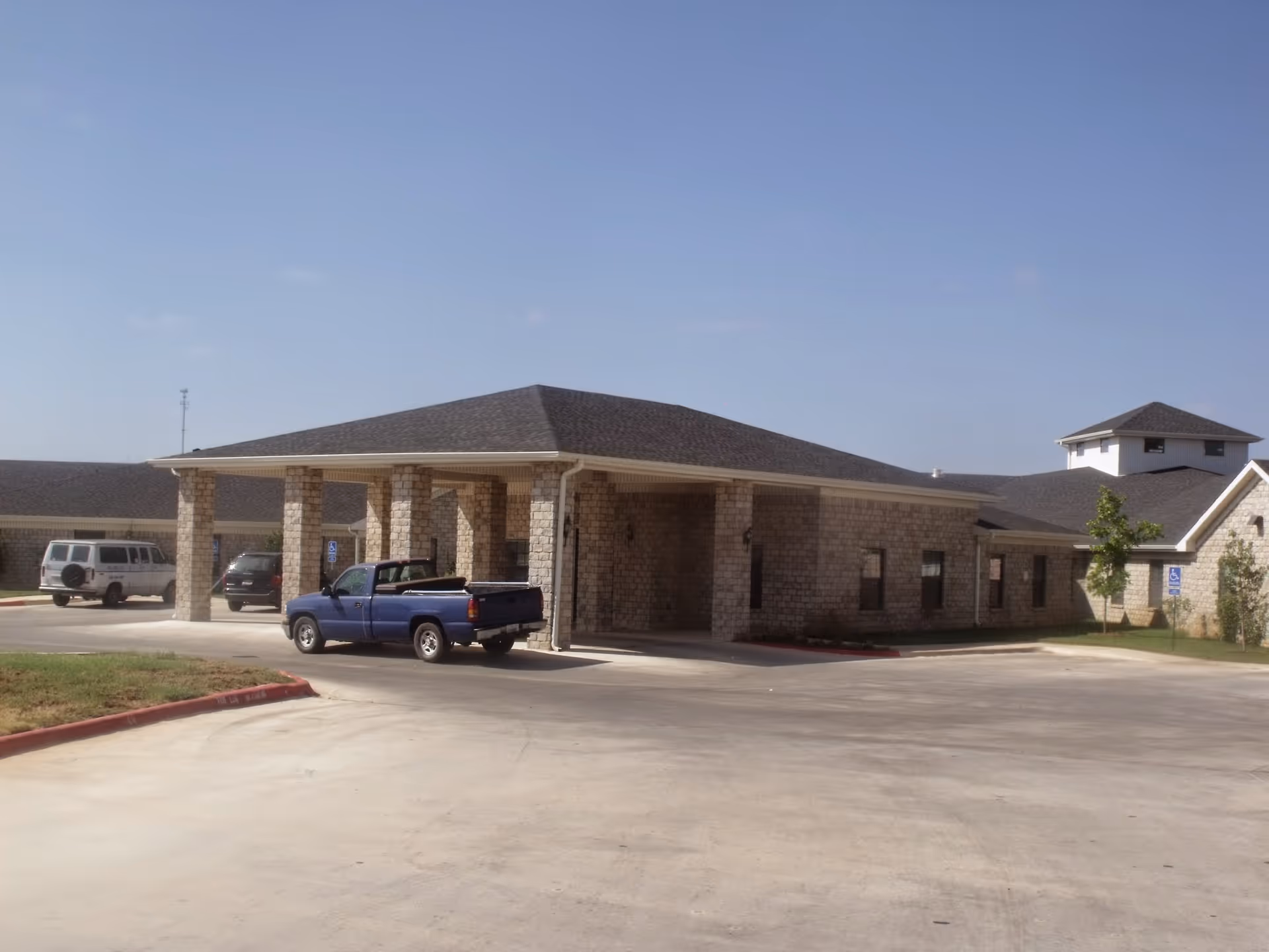Exterior view of Pearsall Nursing and Rehabilitation Center showing a single-story stone building with a covered entrance supported by stone columns. Several vehicles are parked near the entrance under a clear blue sky.
