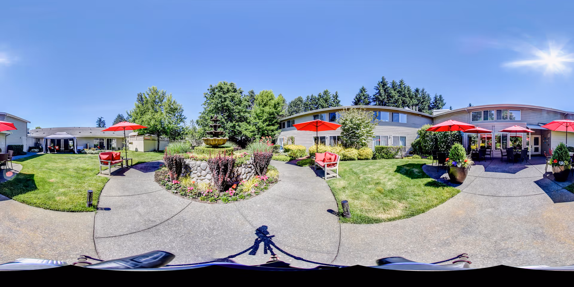 Sunny landscaped courtyard with a central stone fountain, red patio umbrellas, seating, and surrounding two-story senior living building.