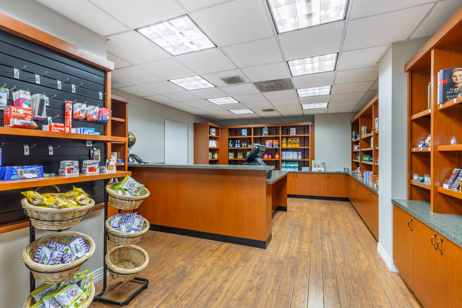 Interior view of a small retail shop area within a facility, featuring wooden shelves stocked with various packaged goods and snacks. There is a wooden counter with a cash register in the center, and the floor is covered with wood-patterned flooring. The ceiling has recessed fluorescent lighting panels.