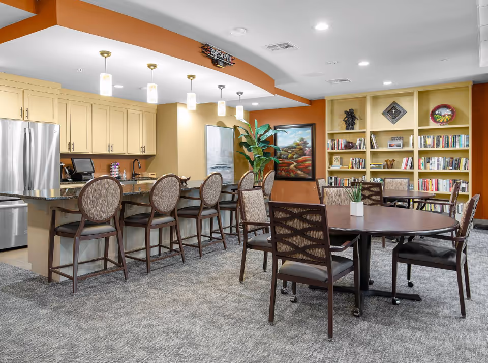 Communal senior living dining and lounge area with a kitchen bar, counter seating, round tables, and bookshelves.